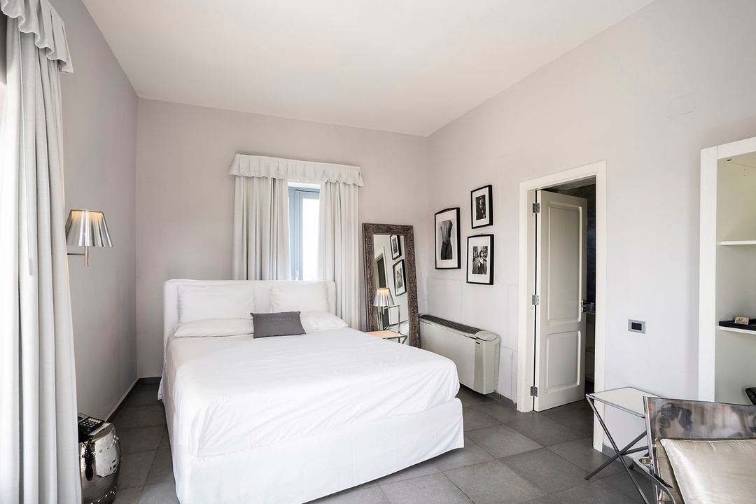 A bright, minimalist bedroom featuring a white bed with gray accent pillow, light purple walls, and modern framed artwork at La Suite Boutique Hotel & Spa.